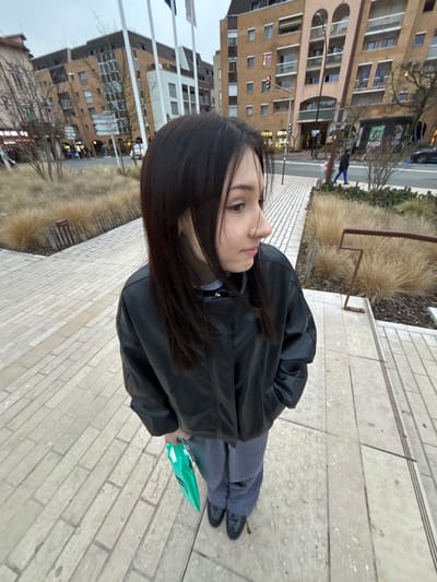 A young woman in a black leather jacket and gray pants stands on a brick sidewalk. She holds a green object in her hand. Buildings line the opposite side of the sidewalk, featuring apartments and storefronts. A patch of tall grass and a metal barrier separate the sidewalk from the street. The sky is overcast.