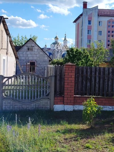 This media file depicts an outdoor scene with several buildings and natural elements. In the foreground, there is a decorative concrete fence with a brick base, separating a grassy area with wildflowers from the background. A small, leafy bush is visible to the right of the fence.
Behind the fence, a house constructed of cinder blocks is partially visible, featuring a simple pitched roof and a window with a damaged pane. Next to this house, on the left, is a portion of another building with a white brick facade and a dark-tiled roof.
Further in the background, a large, multi-story building with a pale yellow and pink facade is prominent. To the left of this building, a domed Orthodox church with golden spires stands against a blue sky with white clouds. The overall impression is of a semi-rural or suburban setting.
The time of day appears to be daytime, indicated by the bright sunlight and the shadows cast on the ground. The weather seems pleasant, with a clear blue sky and scattered clouds. The scene suggests a calm and ordinary day in a place that appears to be Malaryta, Belarus, based on the provided location context. There is no visible text in the image.