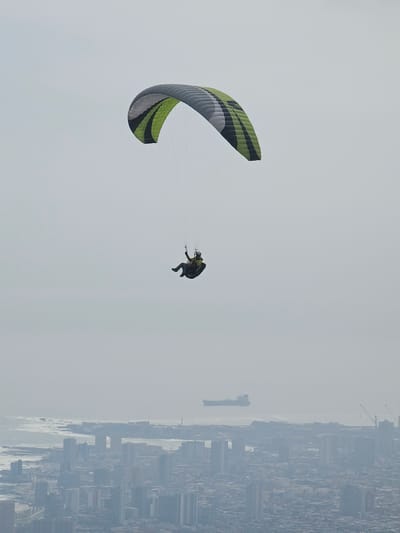 A person paraglides over the city of Alto Hospicio, Chile. The paraglider is striped green, black, and grey. The city is visible in the background, shrouded in a grey haze, with buildings visible. A ship is visible in the water.