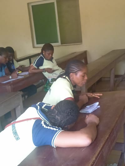 The image depicts a classroom or study hall scene, likely in Nsugbe, Nigeria, showing several young adults or students, primarily female. They are seated at simple wooden desks and benches.

In the immediate foreground, a student wearing a white and blue uniform with "ADVANCED COMPUTING" printed on the back, rests their head down on a wooden desk. Next to them, another student with braided hair is seated, looking towards the right, with several white papers on the desk in front of them.

Further into the background, other students are visible. On the left, one student appears to be writing or focused on materials at their desk, while another student opposite them holds and looks at a mobile phone. A fifth student is partially visible in the far left background.

The room has light-colored walls and features a window with a green, opaque panel in the upper background. The students are wearing uniforms characterized by light-colored (possibly white or cream) shirts with blue sleeves and collar accents. The setting suggests an educational environment during daytime hours, with a generally calm atmosphere.