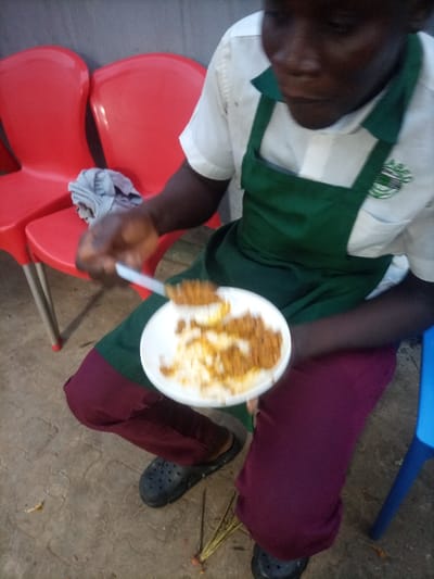 A person wearing a white shirt, green apron, and maroon pants is eating food with a spoon from a white plate. The food appears to be a combination of rice and a meat dish. The person is wearing black Crocs. A red plastic chair with a gray cloth on it sits to the left of the individual, and a blue plastic chair is to the right. Sticks with orange roots are on the ground. The setting seems to be outdoors, possibly a patio or open area.