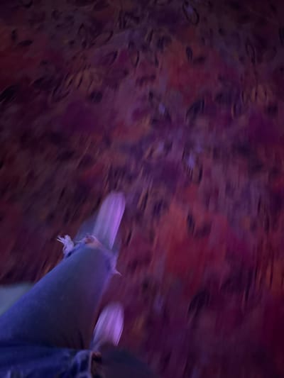 The image captures a close-up, dynamic shot of a person's legs and feet, possibly in motion, against a blurred, abstract background. The background itself is a swirling mosaic of deep purples and reds, with darker, indistinct shapes suggesting a textured or patterned surface. The foreground is dominated by a blue denim-clad leg and a light purple shoe or foot covering, which appears to be made of a soft material, possibly fabric or leather. The shoe has a slightly pointed toe and a frayed edge. The overall impression is one of movement and energy, perhaps a candid shot taken during an activity. The lighting is low and ambient, creating a moody atmosphere. There is no visible text in the image. The context of Haskovo, Bulgaria, does not provide specific visual cues within the image itself, as the focus is entirely on the person's lower extremities and the abstract background.