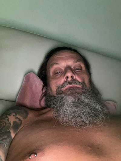 A man with a long, gray beard and long, dark hair is lying down on a light-colored surface, possibly a couch or bed, with a pink pillow supporting his head. He is shirtless and has tattoos on his left shoulder and arm. A silver piercing is visible on his right nipple. The background appears to be a plain, light green wall. The image was taken in Netanya, Israel.