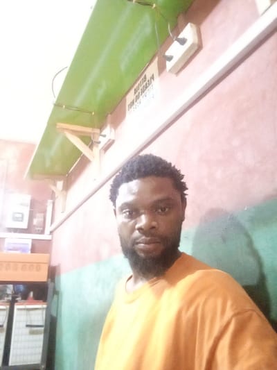 A man with a beard and short, curly hair wears a bright orange t-shirt. He stands inside a room with a color-blocked wall painted pink, then green. A green shelf hangs from the ceiling above him, held up by wooden supports and electrical outlets. A sign is fixed to the wall, illegible due to the angle. A cabinet and batteries are visible on the left side of the frame.
