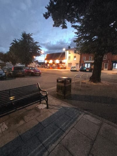 The image depicts a street scene at dusk in Wychavon, United Kingdom. A dark-colored metal bench sits on a paved walkway in the foreground, casting a shadow. To the right of the bench is a dark-colored public trash can with "LITTER" written on it. Across the street, buildings with illuminated windows are visible, including what appears to be a pub with outdoor seating. Several cars are parked or driving on the road, and two U-shaped bicycle racks are positioned near a large tree. The sky is overcast with visible clouds.