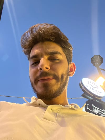 A low-angle, close-up, outdoor selfie shows a young man with dark, wavy hair and a short beard, wearing a white polo shirt. He has dimples and appears to be looking upwards, with the sky in the background. Partially visible in the background are streetlights and signs, including one that reads "STEAM COFFEE SHOP." The lighting suggests it is either dusk or dawn. Based on the presence of the "STEAM COFFEE SHOP" sign, the image is likely located in the Al-Muhajerin Municipality, Syria.