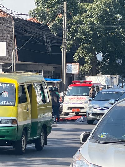 This image captures a scene on a city street in Bandung City, Indonesia, likely near a busy intersection. A yellow and green public minivan, possibly a "angkot," is parked on the left, its door ajar. Beside it, an ambulance with flashing red lights is present, suggesting an emergency situation. In front of the ambulance, a red tarp covers what appears to be a person lying on the ground, implying a possible accident or medical emergency. Several individuals, some wearing helmets and possibly first responders or onlookers, are gathered around the scene. The environment includes buildings with dark facades and tiled roofs, overhead electrical wires, and a large green tree providing shade. The presence of multiple vehicles and pedestrians indicates this is a public thoroughfare.