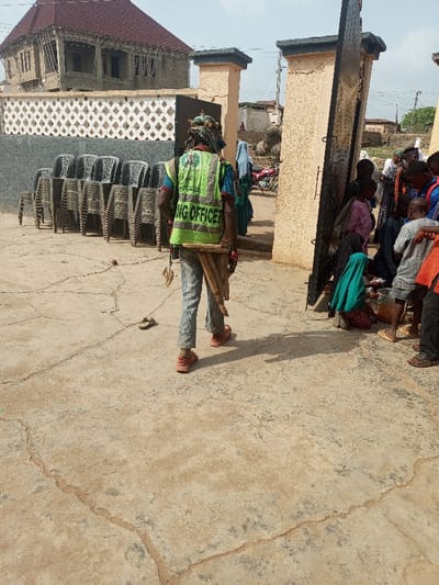 A person wearing a green vest labeled "Polling Officer" walks away from a crowd of people gathered near a stone gate. The person carries a wooden object on their back. To the left, stacked plastic chairs stand against a wall, with a building under construction in the background. The ground is paved and cracked. A flip-flop lies near the person.