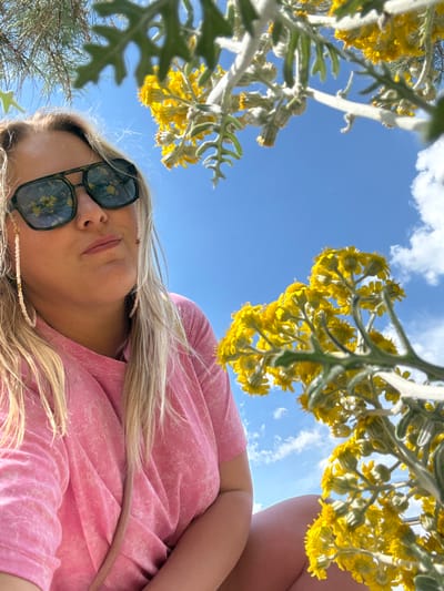 An upward-facing selfie captures a blonde female in Bol'shoi Utrish, Russia, framed by vibrant yellow flowers and a clear blue sky with scattered white clouds. The subject, viewed from the chest up, wears a pink t-shirt with a subtle mottled pattern. She has a neutral expression, with slightly pursed lips. Dark, square-framed sunglasses are worn, reflecting the yellow flowers and blue sky prominently in their lenses. A white beaded chain, with a small star charm on the viewer's right side, is attached to the sunglasses. Her blonde hair falls over her shoulders. The yellow flowers, characterized by dense clusters of small blooms on greyish-green stems with distinct leaves, occupy the upper and right portions of the frame, appearing close to the camera lens. Sparse green foliage is also visible in the upper-left corner, against the sky.
