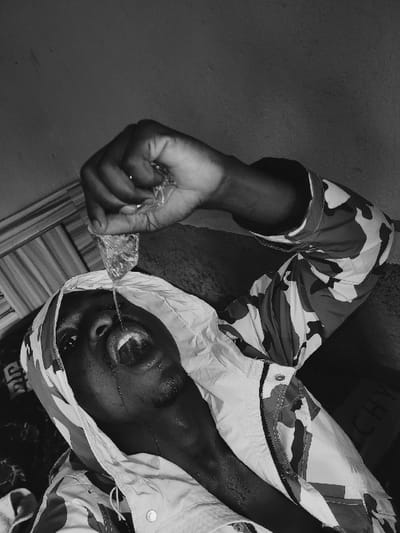 A black and white photograph, captured from a low-angle perspective, depicts a dark-skinned male subject in an indoor setting within Udu, Nigeria. The individual wears a hooded camouflage-patterned jacket, partially unzipped, with the hood covering his head. He is actively pouring liquid from a clear plastic sachet, held in his right hand above his head, directly into his wide-open mouth. His head is tilted sharply backward, and streams of water are visible entering his mouth, spilling down his chin, and wetting his neck. The immediate background shows a plain, textured wall with minor imperfections. A dark surface or fabric is positioned behind his upper back. On the far left, a vertically striped pattern is partially visible, and in the lower right background, the word "GIVENCHY" is faintly discernible on a dark object.