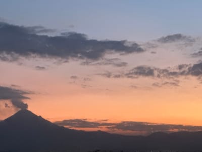 A panoramic shot captures a dramatic volcanic landscape at twilight over what appears to be San Francisco Ocotlán, Mexico. The sky displays a striking gradient of colors, transitioning from a soft blue at the top, through muted purples and pinks, to a warm orange-yellow glow near the horizon. Scattered across the sky are dark, layered clouds, some wispy and high, others denser and lower, partially obscuring the distant mountain ranges to the right.

The prominent subject is a large volcano, silhouetted in dark grey, dominating the left side of the frame. A distinct plume of dark grey smoke or ash billows from its summit, rising upwards and drifting slightly to the left against the colorful sky. Other dark, undulating mountain ranges are visible in silhouette across the lower half of the image. Faint, scattered lights can be discerned at the very bottom edge, suggesting a distant urban area or town nestled at the base of the mountains. The scene depicts a moment of natural activity during either sunrise or sunset. There is no visible text.