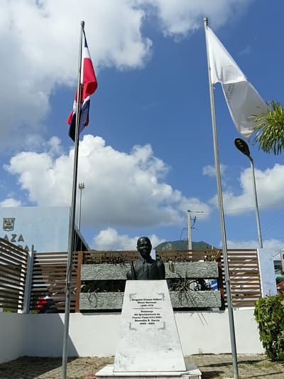 This is a vertical, medium shot of a monument and flags under a bright blue sky with scattered clouds. In the foreground, two flagpoles stand, one flying a white flag with black text and the other flying the Dominican Republic flag. Behind the flags, a bust of a man is visible, positioned in front of a white marble plaque inscribed with text. The plaque is titled "Gregorio Urbano Gilbert Héroe Nacional 1898-1970" and mentions "Homenaje del Ayuntamiento de Puerto Plata 10/01/2021 Diomedes R. Garcia 2020-2024". To the right of the monument is a wooden fence and some greenery. The overall scene suggests a memorial or public space, likely outdoors during the day, given the bright lighting and clear sky. The text indicates the monument is dedicated to a national hero and honors the Ayuntamiento (city council) of Puerto Plata.