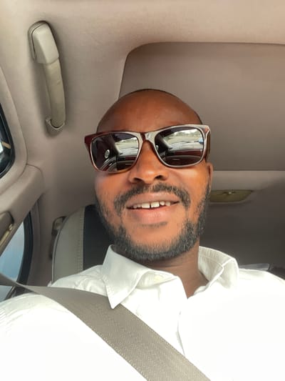 A man wearing sunglasses and a white shirt is in a car. He has a beard and is wearing a seatbelt. The interior of the car is beige. The image does not contain sufficient information to determine the location as Iddo Tudunwada, Nigeria.
