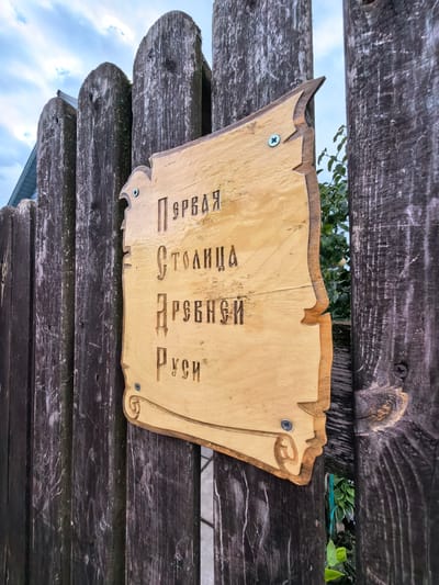 A weathered, light-colored wooden sign with carved Cyrillic lettering is attached to a dark, rustic wooden fence. The sign is shaped like a scroll or a piece of parchment, with decorative edges. The inscription reads "ПЕРВАЯ СТОЛИЦА ДРЕВНЕЙ РУСИ" which translates to "The First Capital of Ancient Rus". The fence is composed of vertical, rough-hewn wooden planks. The background shows a cloudy sky and some green foliage peeking through the gaps in the fence. The location is identified as Staraya Ladoga, Russia, known for its historical significance as the first capital of Rus.