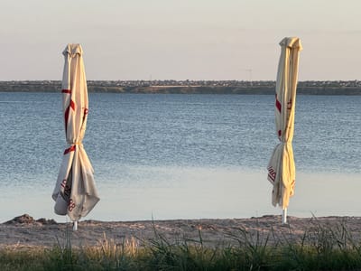 Two closed beach umbrellas stand on a sandy shoreline. The umbrellas are cream-colored with red markings. The one on the left is taller and has a thicker fabric tie around its middle. The one on the right is slightly shorter and has a thinner tie. Both are tied with rope. In the background, a body of water, likely the Dnipro River, stretches to the horizon. Beyond the water, a tree-lined bank is visible, with a distant settlement of houses and buildings. A wind turbine can be seen on the horizon to the right. The sky is a pale blue, transitioning to a lighter tone near the horizon, suggesting either early morning or late evening. The foreground consists of dry grass and sand. This scene is consistent with the shoreline area of Ukrainka, Ukraine, known for its proximity to the Dnipro River. No people are present in the image.