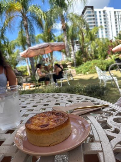 The image captures an outdoor dining scene in East Naples, United States of America, under a clear blue sky. In the immediate foreground, a white, ornate cast-iron table displays a small, round, golden-brown tart on a light pink ceramic plate. A golden-colored fork rests on a light brown paper napkin beside the plate. To the left, a clear glass pitcher, filled with water and showing condensation, is partially visible. A person's upper arm and shoulder, wearing a light-colored strap top, are present in the bottom left corner, suggesting a first-person perspective. The midground, out of focus, shows two individuals seated at a white table beneath a large, peach-colored outdoor umbrella, appearing to converse. One person wears a light-colored top and dark bottoms; the other wears a dark top and dark bottoms. The area around them is a lush green garden with scattered foliage and a white ornate metal chair further to the right. In the distant background, several tall palm trees frame a multi-story, light-colored building with numerous balconies.