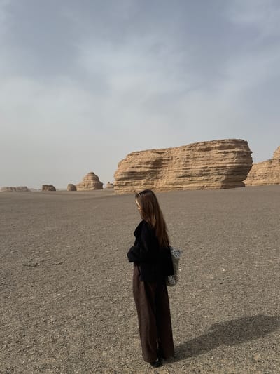 The image captures a landscape in Dunhuang City, China. A person stands on a gravelly surface, wearing a black jacket and brown pants. The background reveals a formation of tall, brown, rock structures under a light grey sky. The overall environment suggests a dry, arid region.