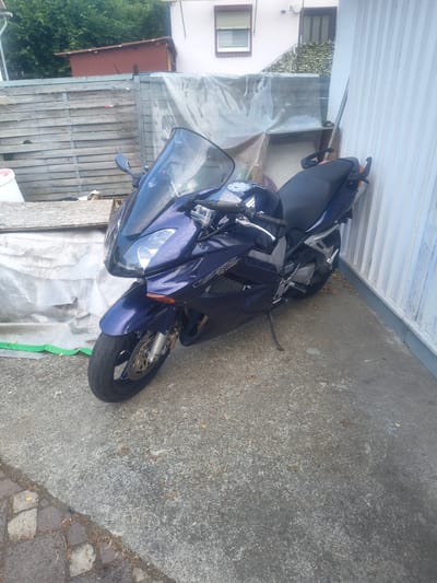 A dark blue motorcycle is parked on a concrete surface next to a white corrugated metal wall. The motorcycle has a clear windshield and a dark seat. To the left of the motorcycle, there is a wooden fence and a large tarp. In the background, there is a building with a window. The ground is covered with some leaves and debris. The scene appears to be an outdoor setting, possibly a yard or driveway. The lighting suggests it is daytime. There is no visible text in the image. The orientation of the image is landscape with the motorcycle running diagonally from the bottom left to the top right.