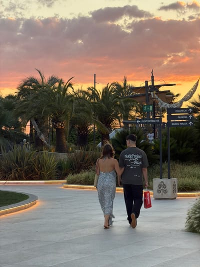 A couple walks hand-in-hand on a paved walkway in what appears to be an outdoor entertainment or shopping area, with a dramatic sunset sky in the background. The woman, on the left, wears a floral-patterned maxi dress and carries a straw bag. The man, on the right, is dressed in a dark t-shirt with graphic text and dark pants, and he carries a red shopping bag. Behind them, palm trees and lush vegetation are illuminated by soft lighting along the walkway's edge. A directional signpost indicates "ASCENSOR," "PARKING," and "BARCO PIRATA" (Elevator, Parking, and Pirate Ship, respectively), and also points to "FUENTE INTERACTIVA" (Interactive Fountain). The overall scene suggests a warm evening, possibly in a resort or themed attraction. Based on the directional signage in Spanish, the location is likely within a Spanish-speaking country. However, there is no specific indication that this scene is located in Majadahonda, Spain.