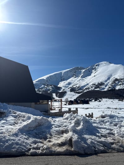 This image depicts a snow-covered mountainous landscape in Virak, Montenegro, under a clear blue sky with intense sunlight emanating from the upper left. In the foreground, a dark asphalt road surface is visible at the bottom, bordered by large, irregular mounds of dirty, partially melted snow. To the left-midground, the corner and side of a modern building with a dark gray, steeply pitched roof and light-colored stone walls are prominent. Adjacent to this building, a stone wall with decorative capstones encloses a small area where a wooden-framed structure supports a large, white fabric hammock or swing. Beyond this, a vast, snow-covered valley extends, dotted with several smaller, dark-roofed wooden cabins. In the background, majestic, snow-capped mountains rise, featuring visible ski slopes and scattered patches of dark, bare trees on their lower flanks. No individuals or specific activities are observable.