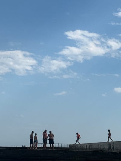 The image depicts a group of young men in swim trunks on the edge of a tall concrete structure, possibly a bridge or a cliff, overlooking a body of water. The sky is a clear blue with scattered white clouds, suggesting a sunny day, likely in the afternoon given the brightness. The men are positioned at different points along the edge, with some standing and others in the process of jumping or about to jump. One individual is in mid-air, appearing to be diving, while another is on the edge, leaning forward. The concrete structure has a railing in some sections. The scene evokes a sense of daring and adventure, characteristic of cliff diving or a similar recreational activity. The overall impression is one of youthful energy and a carefree summer day. There is no visible text in the image.