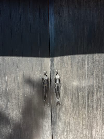 The image shows a weathered grey wooden surface with two decorative metal figurines. The figures appear to be stylized human forms, oriented horizontally. One figurine is on the upper half of the image, and the other is directly below it on the lower half of the image. A stark diagonal line separates a sunlit portion of the wooden surface from a darker, shadowed area. The scene's location is speculated to be in Pecatu, Indonesia, based on contextual information.