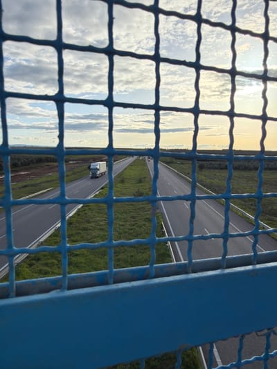The scene is observed from an elevated position, likely an overpass, within Ait Boubidmane, Morocco. A blue metal grid fence, showing signs of paint wear and mild corrosion, occupies the immediate foreground, partially obscuring the view. Below the fence, a solid blue metal beam forms the base of the observation point. Beyond this structure, a modern multi-lane highway extends horizontally across the frame and into the distance, featuring two distinct carriageways separated by a grassy median. The left carriageway shows a white semi-trailer truck traveling away from the viewer. The right carriageway contains multiple smaller vehicles visible in the far distance, traveling towards the viewer. Lush green grass borders both sides of the highway and covers the central median. Beyond the road, the landscape consists of vegetated terrain extending to the horizon, appearing as fields or natural shrubland. The sky above features scattered white clouds, with the sun low on the right side, producing a bright glare. No individuals are present in the observable portion of the scene.