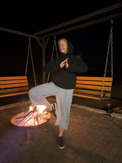 A person is sitting on a hanging bench swing near a fire pit. The person is wearing a black hooded sweatshirt and blue and white striped pants. The fire pit is filled with burning wood. The area around the fire pit is paved with brown bricks. The photograph was taken outdoors at night. The location is Kamennoye, Russia.