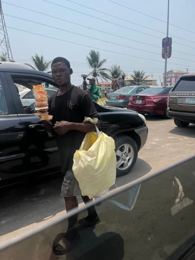 The image depicts a vibrant street scene in Jakande, Lagos State, Nigeria, under bright, clear daylight conditions.
**Central Subject:** An adult male, of dark complexion, stands in the foreground, facing slightly towards the camera. He wears a black short-sleeved t-shirt with a white "GTF" logo on the left chest, grey distressed denim shorts, and dark slides/sandals. His lips are pursed, and his brow is furrowed. He functions as a street hawker, holding a stack of individually packaged snacks (featuring yellow and red packaging) on a stick in his right hand. A large, light-yellow plastic bag, likely containing more merchandise, hangs from his left shoulder. He is positioned alongside the open driver's side door of a stationary dark-colored SUV (possibly a Kia Sportage).
**Midground:** A line of vehicles extends into the background, indicative of traffic. These include a light-green sedan, a red sedan (with a partially visible Nigerian license plate on the rear), and a tan/brown Range Rover SUV displaying a Nigerian license plate "LSD 277HB" on its rear. Beyond the vehicles, several other individuals are visible; one person wears a green jacket and a white cap, and two distinct figures, likely women, are seen carrying items balanced on their heads.
**Background and Environment:** The sky is clear and blue, suggesting fair weather. Palm trees are scattered across the mid-ground, typical of a tropical setting. Overhead power lines crisscross the frame. On the far left, a tall metal lattice power transmission tower is visible. To the right, a utility pole features a purple rectangular advertising banner and a streetlamp. Low-rise buildings are discernible in the distance. Reflections, possibly from a car window, are present in the extreme bottom foreground.