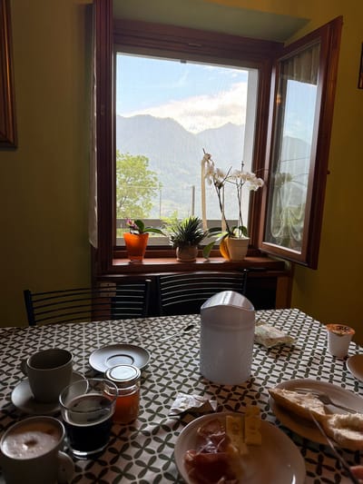 The image depicts a breakfast scene set on a table covered with a green and white patterned tablecloth, positioned in front of a window in Zogno, Italy. Through the window, a mountainous landscape is visible under a sky with clouds. On the windowsill, there are several potted plants, including an orchid with white flowers.
On the table, there are several breakfast items: a white mug with a frothy beverage, a glass of dark liquid, a small jar of honey, and a plate with sliced bread, cheese, and cured meat. A white cylindrical object, possibly a sugar dispenser or a napkin holder, is also on the table, along with a small cup of yogurt. Two chairs with metal frames are visible behind the table. The room has olive green walls. The overall ambiance suggests a peaceful morning meal with a view of the Italian countryside.