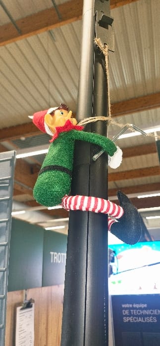 An elf-like plush toy is prominently displayed, hanging from a dark cylindrical structure in an indoor commercial or industrial setting in Vendin-le-Vieil, France. The toy is predominantly green, featuring a red pointed hat, pointed ears, and a light-colored face. Its legs are distinctively striped in red and white.
The activity depicted is the elf toy "climbing" or "hanging" from the cylindrical object. Its arms are wrapped around the cylinder, with one arm appearing to grasp a small, reflective metal hook or piece. Its striped legs are also curled around the cylinder, reinforcing the impression of playful suspension.
Notable details include a piece of brown twine securely tied around the cylindrical object near the elf's head, and a metal bracket situated close to the twine. The background consists of a light-colored, corrugated metal ceiling and visible wooden support beams, suggesting a functional space like a warehouse or workshop. In the deeper background, a blue screen or poster is visible, displaying French text that reads: "votre équipe de TECHNICIENS SPÉCIALISÉS" (your team of SPECIALIZED TECHNICIANS). Below this, a partially visible green object has the word "TROT" printed on it, and another lighter object, possibly a clipboard, is also present. The overall scene, captured in Vendin-le-Vieil, France, integrates a whimsical decoration within an otherwise practical environment.