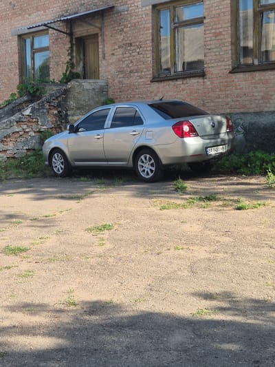 The image shows a silver sedan parked on a dirt and gravel road next to a weathered brick building. The building has several windows, some with boarded-up panes, and a wooden door. Greenery, including vines, grows around the base of the building and along the edge of the road. The car's license plate is visible, reading "BA 8451 HB". The scene appears to be set outdoors, with dappled sunlight suggesting it is daytime. The overall impression is one of a quiet, somewhat neglected urban or semi-rural setting. The image is oriented vertically, with the car and building occupying the left side and the road taking up the right.