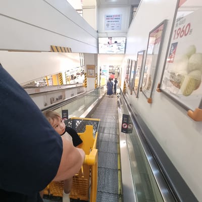The image shows an indoor escalator in a retail environment, likely a department store or supermarket, given the presence of promotional displays and product information on the walls. The escalator is moving, and a young child is seated in a yellow shopping cart next to the camera, looking to the right. The child is wearing a black and white shirt and white socks. In the background, several adults are visible walking on the escalator. One adult is wearing a blue dress, and another is wearing black clothing. Promotional signs are displayed on the walls alongside the escalator, with one sign showing a price of "1,960". Another sign appears to be an advertisement for "e•mart". The escalator is enclosed by glass railings. The overall environment suggests a commercial space with a focus on shopping. The mention of "e•mart" suggests the location is a branch of the E-Mart retail chain, which has numerous locations throughout South Korea, including in Wonju-si.