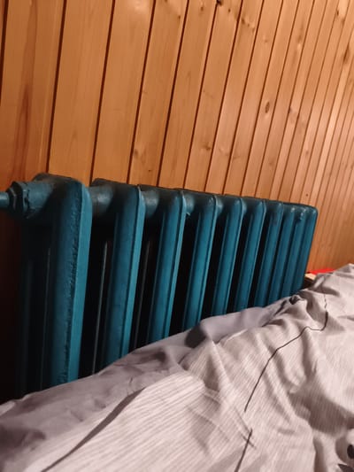 The image shows a teal-colored cast-iron radiator against a background of vertical wooden paneling. In the foreground, a portion of a bed with patterned bedding is visible. The scene appears to be an indoor setting, possibly a room in a home. There are no people or visible actions depicted. The lighting suggests it may be evening or a dimly lit room. The overall impression is one of a domestic interior. There is no information within the image to suggest its location in Sputnik, Russia.