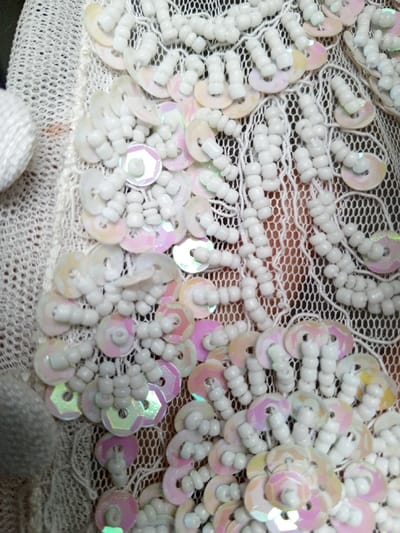 An intricate design features shimmering, iridescent sequins arranged in floral patterns interspersed with delicate, netted lace. White, cylindrical beads accentuate the sequin embellishments, creating raised textures. Some fabric or detailing is also visible on the left. A delicate and textured surface results from the combination of sequins, beads, and lace.
