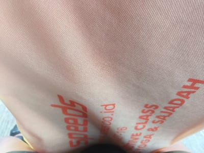 A close-up shot of a light orange, textured fabric with red lettering. The word "speeds" is visible on the lower left, while a website address ending in ".co.id" appears near the center. Further right, the phrase "TIVE CLASS YOGA & SAJADAH" is stacked vertically. The fabric seems to be part of a bag or item of clothing. The lower portion of the image shows a glimpse of what appears to be a leg, adding context to the image's perspective.