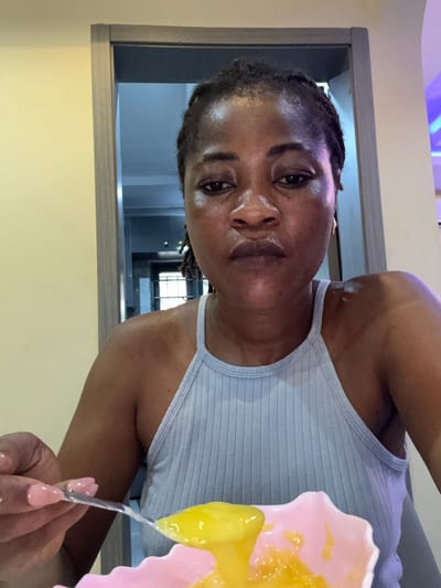 An adult Black woman is captured in a medium, head-and-shoulders selfie. She is wearing a light blue, ribbed tank top. Her braided hair is dark and pulled back. She has brown eyes, and her facial expression is neutral. The woman is holding a spoon with yellow food and is about to eat from a pink bowl. Her fingernails are painted a light pink. The background suggests an indoor setting, possibly a home, with a doorway and part of a wall visible. The lighting is somewhat dim, with some reflections on her skin, indicating perspiration. The scene appears to be located in Ajara, Nigeria.