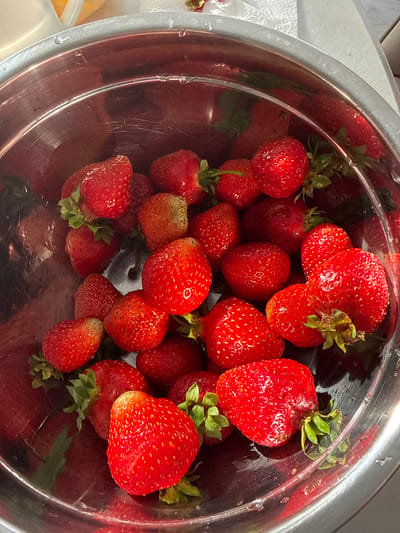 A close-up, overhead shot shows a metallic bowl overflowing with ripe strawberries. The strawberries are a vibrant red with visible seeds and green stems. Some strawberries are clustered together, while others are scattered throughout the bowl. The metallic bowl has a slightly reflective surface, showing some water droplets and subtle scratches. The lighting appears to be natural, suggesting it could be daytime. The background is blurred but hints at a white surface, possibly a countertop or a towel, and a sliver of a white paper towel. The scene evokes a feeling of freshness and abundance, likely in a kitchen setting. There is no visible text in the image. The context provided is "Нижнеяркеево, Russia".