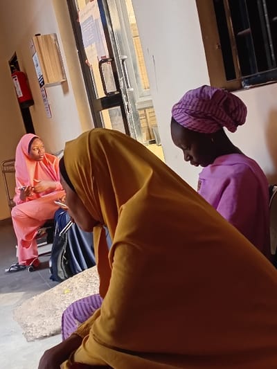 Three women are pictured in a room. One wears a pink hijab, holding an object. Another wears a purple head covering and a light purple outfit. The third woman is wearing a mustard yellow outfit. A window is visible, and a red fire extinguisher is also present.