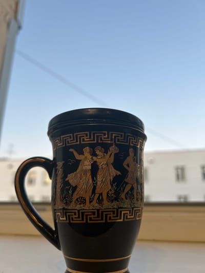 An ornate black mug with gold detailing sits on a windowsill, with a bright blue sky and a blurred cityscape visible in the background. The mug features classical figures in gold, reminiscent of ancient Greek art, and is adorned with a Greek key border. A window frame is visible on the left side of the image.