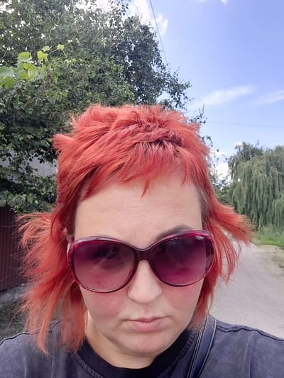 The image is a close-up, portrait-style selfie of a person with vibrant, reddish-orange hair. They are wearing large, dark red sunglasses. The person's face is angled, with their lips slightly parted. They are wearing a dark-colored garment, possibly a jacket or shirt. The background is blurred but suggests an outdoor setting with green foliage and a bright blue sky with some white clouds. A power line is visible against the sky. The overall impression is casual, perhaps taken during a sunny day. The lighting suggests it's daytime. There are no visible signs or text in the image. The style of the photograph, particularly the angle and focus on the person's face and hair, suggests it might be a candid or artistic self-portrait. The background foliage and the clear sky hint at a natural or semi-rural environment, consistent with the Samar, Ukraine location.