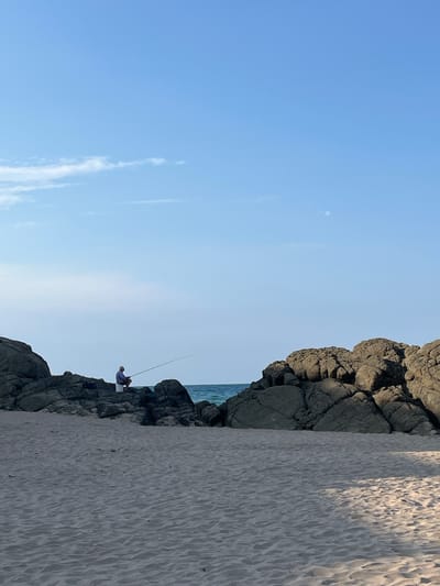 The image depicts a serene beach scene in Ahtopol, Bulgaria. In the foreground, a sandy beach stretches across the frame, showing gentle undulations from the wind or tides. Dominating the midground are large, grey, rugged rocks, forming a natural barrier between the beach and the sea. The rocks are textured and weathered, with visible crevices and shadows.
Between the rocky outcrops, a sliver of the calm, turquoise sea is visible, meeting a hazy horizon. A single individual, a man, is seated on the rocks on the left side of the image. He is wearing a light-colored shirt and appears to be fishing, holding a long fishing rod that extends out over the water. Next to him sits a white bucket.
The sky above is a clear, pale blue, with a few wispy white clouds scattered towards the left. The lighting suggests it is either early morning or late afternoon, with a soft, diffused sunlight casting subtle shadows. The overall atmosphere is one of peaceful solitude and a connection with nature.