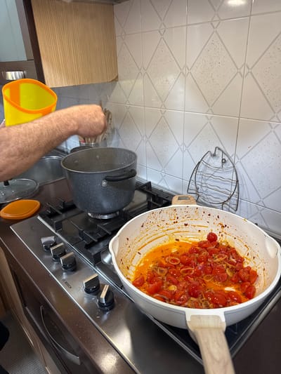 A kitchen scene shows a gas stovetop with two pots, one with a lid nearby. A large white pan sits on the right burner, containing what appears to be a tomato-based sauce. A person's arm and hand are visible, holding a yellow and orange pitcher, potentially pouring liquid into the pot on the left burner. The stovetop is stainless steel and has "Bosch" branding. The backsplash features patterned white tiles, and a fish-shaped decoration hangs on the wall above the stovetop. The cabinets are light brown. This kitchen is potentially located in a residence in Aprilia, Italy.