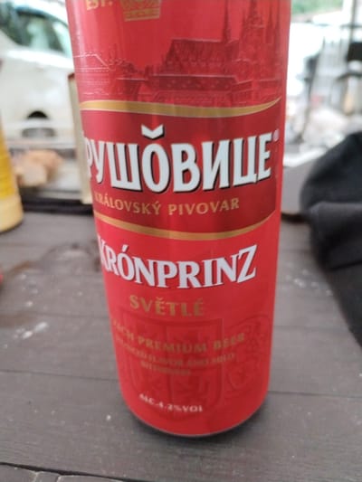 A close-up, vertical shot shows a red beverage can, likely beer, with white and gold text and graphics. The can is the primary subject, positioned centrally and taking up most of the frame. The background is blurred, suggesting an outdoor setting with hints of a table or countertop and possibly other objects or a natural environment.

The can features several lines of text, some in Cyrillic script and some in Latin script. Prominently displayed is "РУШОВИЦЕ" in large white Cyrillic letters with a gold outline. Below this, "KRÁLOVSKÝ PIVOVAR" is written in smaller gold Latin letters. Further down, "TRÓNPRINZ" appears in large white letters with a gold outline, followed by "SVĚTLÉ" in gold. Smaller text below this includes "ACH PREMIUM BEER", "UNCED FLAVOR AND MILD", and "BITTERNE". At the very bottom, "MC.4.2%VOL" indicates the alcohol by volume.

The text suggests the beverage is a beer from a "Royal Brewery" with the name "Ruzovice" and "Kronprinz" as a type or brand. The overall impression is one of a casual setting, perhaps a picnic or outdoor gathering. There are no people visible in the frame. The lighting suggests it could be daytime, with natural light illuminating the can. The surface beneath the can appears to be a textured wooden table.