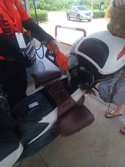 A gas station attendant in Plai Phraya, Thailand, is filling the fuel tank of a white scooter. The attendant wears a red shirt with black sleeves and a lanyard with an ID. The scooter is fitted with brown leather saddlebags and other camping gear. A passenger wearing sandals stands nearby. A silver pickup truck is parked in the background, partially obscured by green shrubbery.