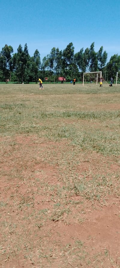 The image shows a dirt field with sparse grass, likely used as a soccer field, in Kipkarren, Kenya (latitude: 0.613, longitude: 34.975).  There are five people on the field, most of whom are wearing yellow shirts. One person is in the middle of the field, holding a soccer ball and appears to be getting ready to take a shot. There is a soccer goal at the back of the field. The ground is red-brown dirt with patchy, sparse, green grass. In the background there is a line of trees, a clear blue sky, and several red-roofed structures.
