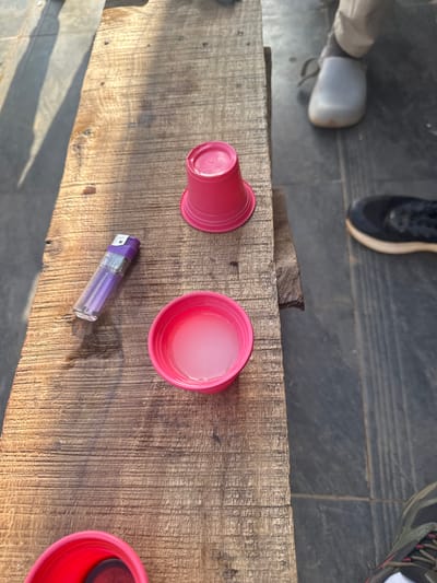 The image shows a weathered wooden surface, possibly a bench or table, with several items on it. There are three red plastic cups, one overturned, one filled with a translucent liquid, and one partially visible at the edge of the frame. A clear plastic lighter with a purple top lies between two of the cups. Parts of footwear belonging to unseen people are visible at the top of the image. The image was taken in Kurunduma, Nigeria.