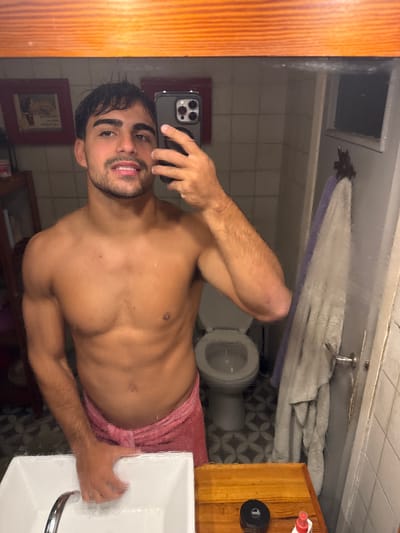 A shirtless man is taking a mirror selfie in a bathroom. He is holding a smartphone with a triple-lens camera. He is wearing a pink towel. A toilet, a towel rack, and a sink are visible in the background. Tile patterns are on the bathroom floor and walls. A wooden cabinet is on the right of the frame. There is also framed art on the wall. There is no indication that the photo was taken in Avellaneda, Argentina.