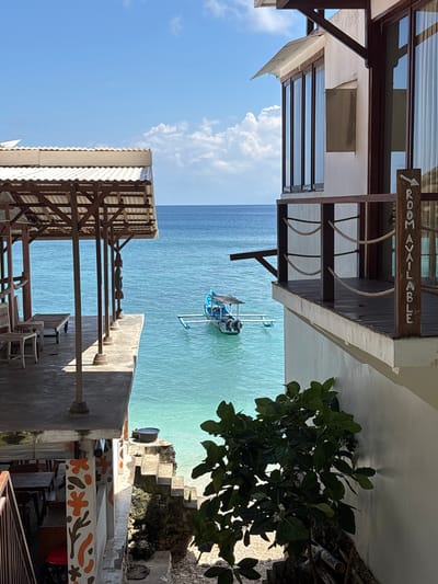The image captures an ocean view in Uluwatu, Indonesia, likely taken from an elevated position, possibly a hotel or guesthouse. The perspective is somewhat tilted. The foreground features architectural elements of the building: wooden beams, a white awning, and sections of walls. A partially obscured sign reads "ROOM AVAILABLE" (the spelling is slightly altered). A lush green tree is on the right side.
In the middle ground, a small boat with a blue and white canopy is moored in the clear turquoise water. Beyond the boat, the ocean extends to the horizon.
The sky is blue, with a few white clouds visible to the left of the frame. The photograph appears to be taken during the day, with natural sunlight illuminating the scene. There are no people visible in the image.