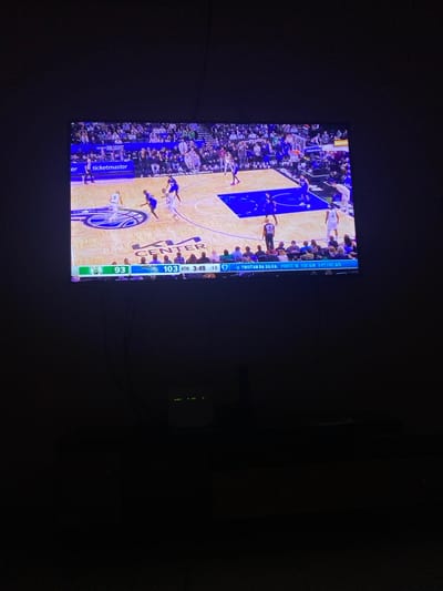 The media file presents a television screen displaying a basketball game in a very dark room, possibly at night, in Lakka, Sierra Leone. The screen is the primary light source, illuminating the basketball court and players, while the surrounding environment remains obscured. To the right of the television, a small electronic device with three distinct green indicator lights is visible against the dark background.

The TV screen shows an active basketball game with multiple players on the court, dressed in blue and white uniforms. The court itself is made of light-colored wood, featuring a large circular "CENTER" logo in the middle and a "ticketmaster" advertisement along the sideline.

A detailed scoreboard and game information are visible on the bottom right of the screen. One team has scored 103 points, while the opposing team has 93 points. The game is in the "4TH" quarter, with "3:49" minutes remaining on the game clock and ":16" seconds on what appears to be a shot clock. Individual player statistics are also displayed, showing "TRISTAN DA SILVA" with "POINTS 15," "FGS 5/8," and "3-PTIGS 3/5." The team name "MAGIC" is also visible. The overall scene captures the experience of watching a live sports broadcast in a darkened setting.